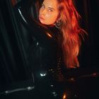 Keine sichtbaren Texte. Eine Person in schwarzem glänzendem Outfit, mit rotem Licht akzentuiert und Hand im Haar.