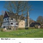 März / April 2027, 12./13. Woche. Haus am Quall in Haan-Gruiten. Fachwerkhäuser mit Kirchturm vor grünem Feld und Bäumen.