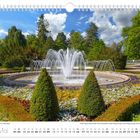 Mai, Springbrunnen im Albertpark, umgeben von bunten Blumenbeeten, mit blühenden Bäumen und bewölktem Himmel im Hintergrund.