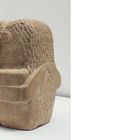 Quadratische Skulptur mit stilisierter Frisur und gekreuzten Armen, aus rauem, sandfarbenem Material.