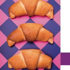 Drei goldbraune Croissants auf einem blau-pinken Rautenmuster. Keine Texte enthalten.