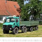 Auf einer Wiese steht ein grüner Unimog mit Anhänger vor einem Fachwerk- und einem Ziegelhaus mit Solarpanel.