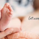 Text: "Gott wird Kind!" Zwei Babyfüße ruhen sanft in einer Hand, umgeben von weicher, warmer Decke.