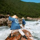 Zwei Personen in blauen Jacken mit Symbolen navigieren ein Floß durch einen wilden Fluss, umgeben von grünen bewaldeten Hügeln.