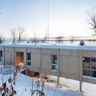 Keine sichtbaren Texte. Ein modernes, rechteckiges Haus auf Stelzen in verschneiter Landschaft, Bäume im Hintergrund.