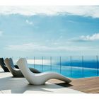 Keine Texte erkennbar. Drei moderne Liegestühle neben einem Pool mit Blick auf das Meer unter blauem Himmel.
