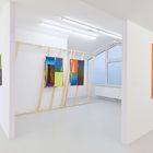 Helle Galerie mit abstrakten, farbenfrohen Gemälden an weißen Wänden und einer Holzkonstruktion. Große Fenster im Hintergrund.