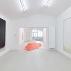 Ein heller Raum mit abstrakten Gemälden an den Wänden und einem rot-orangenem, sternförmigen Teppich auf dem Boden.