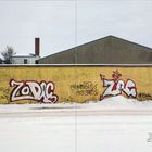 Graffiti auf gelber Wand mit den Worten "ZODIAC", "COLD CRUSH", "ZAC", umgeben von Schnee und Gebäuden im Hintergrund.