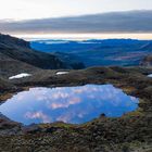 Keine Texte im Bild. Eine beeindruckende Berglandschaft mit spiegelndem See und Hügeln unter einem bewölkten Himmel zur Dämmerung.