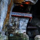Steinige Höhlenwand mit zwei überdachten Brücken und ruhigem Wasser darunter. Oben ein oranger Lichtschein.
