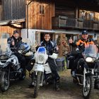 Drei Personen in Motorradkleidung sitzen auf Motorrädern und zeigen Daumen hoch, vor einem rustikalen Holzhaus.