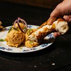 Eine Hand taucht eine Brezel in orangefarbenen Aufstrich auf einem Teller mit Blumenmotiv, garniert mit Zwiebeln.