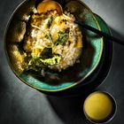 Eine grüne Schale mit Risotto, Mais, Blättern und einem goldenen Löffel. Neben der Schale eine Kelle mit gelber Flüssigkeit.