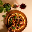 Keine Texte vorhanden. Eine Pizza mit Basilikum, Oliven und Pilzen auf Holzbrett. Hand nimmt ein Stück. Basilikum und Rotweinglas daneben.