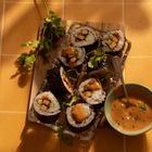 Sushis mit Tofufüllung auf Holzbrett, daneben eine grüne Schale mit Soße; Korianderblätter dekorieren alles.