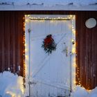 Weiße Holztür mit einem Kranz und roter Schleife dekoriert, umrahmt von warmen Lichterketten im verschneiten Umfeld.