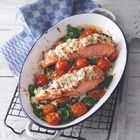 Zwei Lachsfilets mit Käse überbacken, umgeben von Kirschtomaten und grünem Gemüse in einer ovalen Form. Daneben ein blau-weißes Tuch.