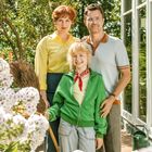 Keine erkennbaren Texte. Drei Personen lächeln im Garten, umgeben von weißen Blüten. Hinten ein Mann und eine Frau.