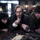 Drei Personen liegen am Boden in einem Raum, schauen intensiv nach vorne. Die Szene wirkt geheimnisvoll und spannend.