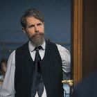 Ein Mann mit Bart in eleganter Kleidung steht nachdenklich an einer Tür und schaut auf eine andere Person.