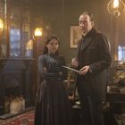 Zwei Personen in einem viktorianisch eingerichteten Raum mit warmem Licht. Eine Frau steht mit verschränkten Armen. Ein Mann hält ein Buch.