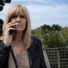 Eine Frau mit blonden Haaren telefoniert draußen. Sie trägt eine schwarze Lederjacke. Im Hintergrund sind Bäume und Hügel.