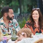 Ein Mann und eine Frau sitzen am Tisch im Freien, umgeben von Essensgeschirr, während ein Kind vorne spielt.