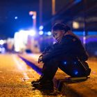 Eine Person in Uniform sitzt nachdenklich an einem Bordstein, umgeben von unscharfem, blauem Licht im Hintergrund.