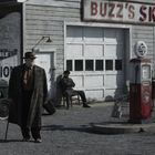 „Buzz's Skelly“ auf einer Tankstelle, darunter alte Zapfsäulen. Ein Mann mit Hut und Mantel geht am Gebäude vorbei.