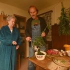 Ein Mann zerkleinert Pflanzen mit einer alten Mühle, eine ältere Frau beobachtet. Kräuter hängen von der Decke.