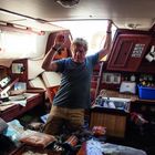 Ein Mann in einem überfluteten Boot hält sich an der Decke fest, umgeben von schwimmenden Gegenständen.