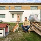 Ein Mann in gelbem Hemd sitzt neben einem Hund vor einem weißen Haus mit Baustellenmaterial und einem Hundehäuschen im Vorgarten.