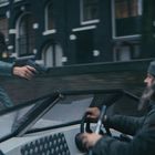 Eine Person auf einem Boot zielt mit einer Pistole auf den Fahrer, der warm angezogen ist. Gebäude im Hintergrund.