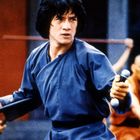 Ein Mann in blauer Kleidung hält zwei Stöcke. Im Hintergrund üben Menschen mit Stäben Kampfkunst.