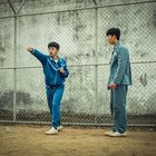 Zwei Männer in Trainingsanzügen stehen vor einem Maschendrahtzaun, einer übt einen Kampfsportkick, während der andere zusieht.