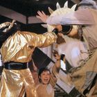 Drei Personen in Bewegung, eine in goldener Kleidung, eine trägt ein rundes Schild. Alle scheinen in einer Kampfszene zu sein.