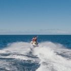 Zwei Personen fahren auf einem Jetski über das offene Meer, hinterlassen spritzende Gischt. Klarer blauer Himmel.