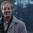 Ein lächelnder Mann mit Brille und Schnurrbart steht draußen in einem Herbstmantel, Hintergrund unscharf.