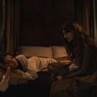 Zwei Frauen in einem dunklen Raum. Eine liegt auf einem Bett, die andere sitzt und spricht. Kerzenlicht erhellt die Szene.