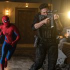 Ein Mann in Spiderman-Kostüm läuft hinter einem bewaffneten Mann her, der ein Gewehr abfeuert. Die Szene wirkt dynamisch.