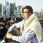 Ein Mann in einem Pullover sitzt am Wasser, mit einem Hafen voller Boote und hohen Gebäuden im Hintergrund.