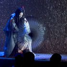 Eine Figur in traditioneller Kleidung hält einen Regenschirm im Regen. Publikum im Vordergrund, beleuchtet von Bühnenlicht.