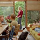 Ein gemütlicher Raum: Kinder am Tisch, eine Frau draußen, ein Hund am Boden, ein Kind liegt auf dem Sofa.