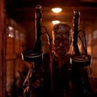 Eine schmutzige Person steht in einem dunklen, hölzernen Raum, hält zwei Maschinenpistolen.