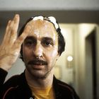 Ein Mann mit schütterem Haar und Schnurrbart, Gesicht mit Ei übergossen, hebt eine Hand hoch, in einem hellen Raum.