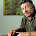 Ein nachdenklicher Mann mit Bart in einem olivgrünen Hemd sitzt an einem Tisch, im Hintergrund unscharfe grüne Formen.