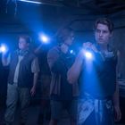 Drei Personen in dunklem Raum mit Taschenlampen, tragen Schutzmasken um den Hals. Sie wirken aufmerksam und angespannt.