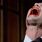 Ein Mann mit gebleckten Zähnen, blutigem Mund, Kopf im Nacken, trägt Smoking, vor dunklem Hintergrund.