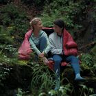 Ein Mann und eine Frau sitzen gemeinsam in einem roten Schlafsack auf einem moosigen Felsen im Wald und sehen sich an.